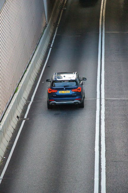 An overhead view of a light-colored passenger car driving along a multi-lane urban road with clearly marked dashed white lane lines. The road is relatively empty, with no other vehicles visible in the