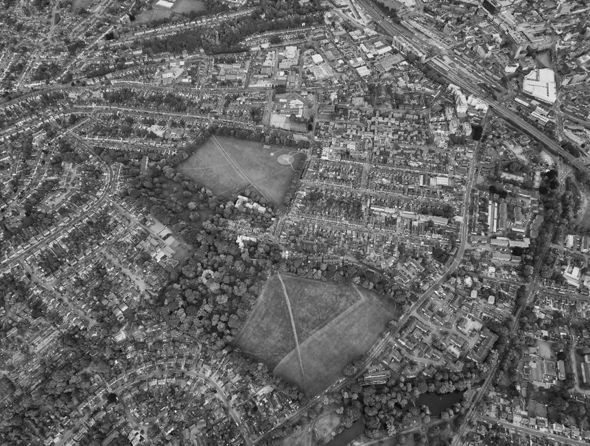 Aerial black-and-white photograph of a residential neighbourhood showing dense housing and streets. Near the center, there is a large park with two open grassy areas separated by a pathway, surrounded