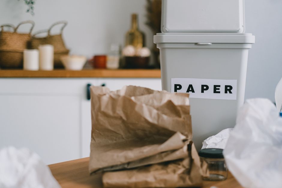 A person's hand holding a white plastic bag, positioned above a white bowl filled with partially decomposed food scraps, including vegetable peels and food waste, placed on a green plastic container b