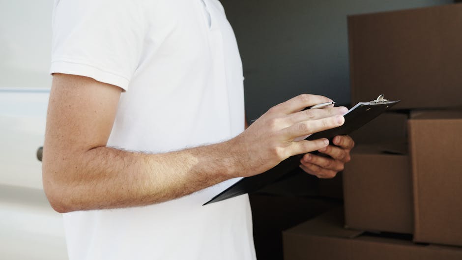 Close-up of a person wearing a white t-shirt, holding a black clipboard and pen, inspecting and making notes during a home relocation process. In the background, several cardboard moving boxes of vari