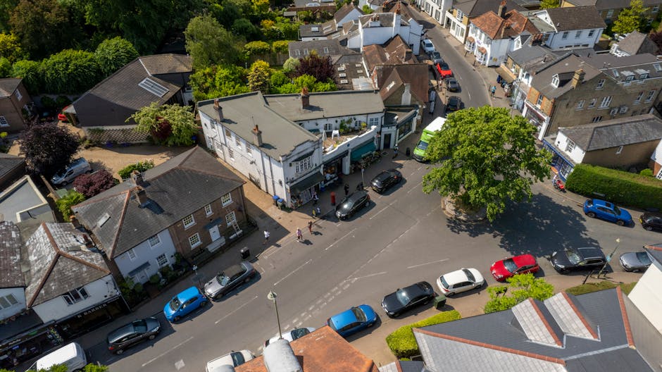 An aerial view of a residential street showing a house being loaded with items for a home relocation, with a green van parked nearby and several black, white, red, and blue cars parked along the pavement. The house, situated at the corner of the street, has a white exterior with a small porch area and a flat roof. Moving blankets and packing material are visible around the entrance, indicating the packing and furniture transport process. A large tree provides shade in the vicinity, with additional houses and greenery lining the street in the background. The scene captures the loading process conducted by Man With a Van Pinner, highlighting typical elements of local removals and the organization involved in house moves within the community.