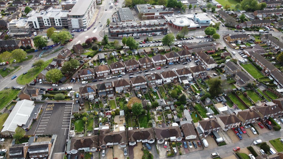 An aerial view of a residential street showing a house being loaded with items for a home relocation, with a green van parked nearby and several black, white, red, and blue cars parked along the pavement. The house, situated at the corner of the street, has a white exterior with a small porch area and a flat roof. Moving blankets and packing material are visible around the entrance, indicating the packing and furniture transport process. A large tree provides shade in the vicinity, with additional houses and greenery lining the street in the background. The scene captures the loading process conducted by Man With a Van Pinner, highlighting typical elements of local removals and the organization involved in house moves within the community.