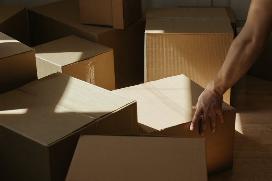 A person’s hand is seen placing a cardboard box onto a stack of similar boxes inside a room with natural light casting shadows. The boxes are made of brown corrugated cardboard and are arranged on a hardwood floor, some stacked on top of each other while others are lined up. The scene suggests a home relocation or packing process, with the individual preparing for furniture transport and moving. In the background, additional boxes are visible, indicating an organized packing session typical of house removals. The lighting highlights the textures of the cardboard and the wooden flooring, and the person’s arm and hand are partially visible as they handle the box carefully. This image depicts the careful packing and loading process involved in household relocations, with the focus on box handling and space organization, aligned with the services offered by Man With a Van Pinner for small flat moves near Pinner station, including packing and moving logistics.