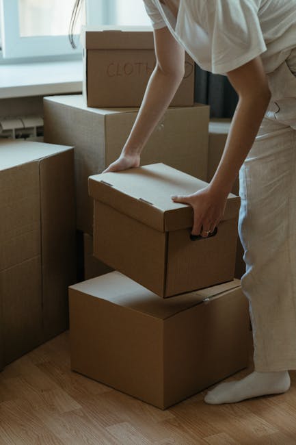 A person’s hand is seen placing a cardboard box onto a stack of similar boxes inside a room with natural light casting shadows. The boxes are made of brown corrugated cardboard and are arranged on a hardwood floor, some stacked on top of each other while others are lined up. The scene suggests a home relocation or packing process, with the individual preparing for furniture transport and moving. In the background, additional boxes are visible, indicating an organized packing session typical of house removals. The lighting highlights the textures of the cardboard and the wooden flooring, and the person’s arm and hand are partially visible as they handle the box carefully. This image depicts the careful packing and loading process involved in household relocations, with the focus on box handling and space organization, aligned with the services offered by Man With a Van Pinner for small flat moves near Pinner station, including packing and moving logistics.